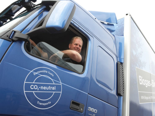 Bilde av mannlig sjåfør av blå lastebil. Lastebilen har et merke som tilsier at bilen bruker fornybart drivstoff og er CO2 nøytral.