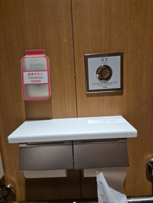 bildet viser en toalettrull-holder som har innebygget hylle på oversiden hvor det er mulig å legge fra seg utstyr. Det er også bilde av en alarmknapp for assistanse som er montert like ved rullen.