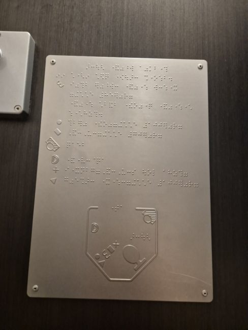 bildet viser et taktilt skilt på et tog-toalett. Skiltet gjør det mulig å få oversikt over rommet uten å måtte føle seg fram med hendene da både vask, toalett, dør og toalettruller er markert med taktile symboler.