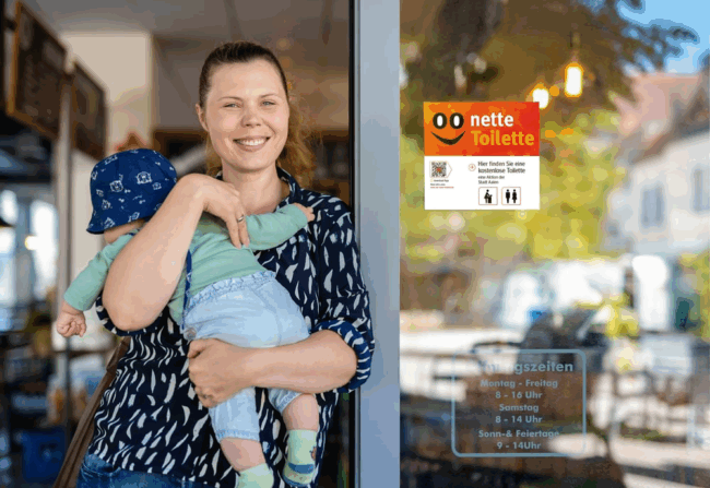 Bildet viser en dame og en baby som står i en døråpning på en kafe. Det henger et skilt på kafeen hvor det står «Nette toilette» for å vise at kafeen er med i ordningen.
